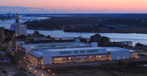 Aerial view of the Wilmington Convention Center at twilight along the river, with illuminated windows, nearby streets, and a bridge visible in the distance beneath a fading sunset sky.