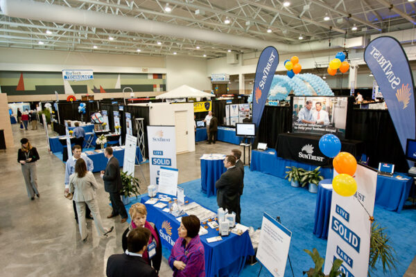 Wide view of a convention hall trade show with branded booths, blue carpeted display areas, balloons, and attendees walking and talking across the exhibit floor.