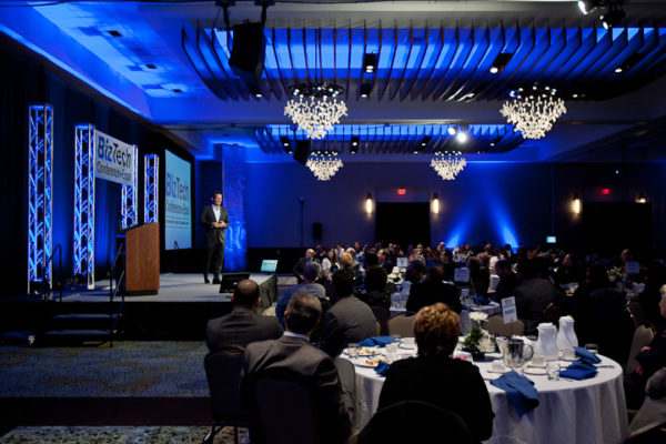 Audience seated at round banquet tables in a dimly lit event ballroom while a speaker presents on stage under blue lighting and chandeliers.