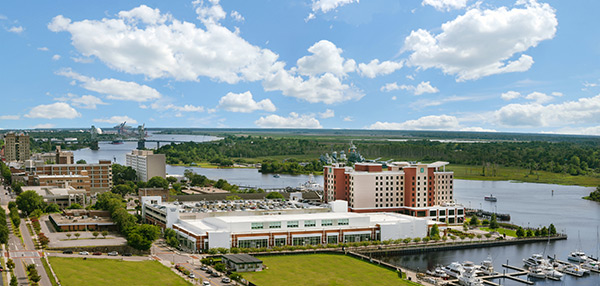 Aerial view of the Wilmington Convention Center along the river, with nearby hotels, marina docks, surrounding city blocks, and a broad expanse of water under a partly cloudy sky.