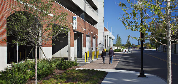 Street-level view of a parking structure entrance beside a wide sidewalk with trees, landscaping, and two people walking on a clear day.