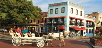 Horse-drawn carriage carrying passengers along a brick street in front of a two-story corner building with red awnings and nearby trees.