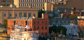 Soft-focus view of downtown buildings in warm light, including a white riverboat in the foreground and brick buildings behind it.