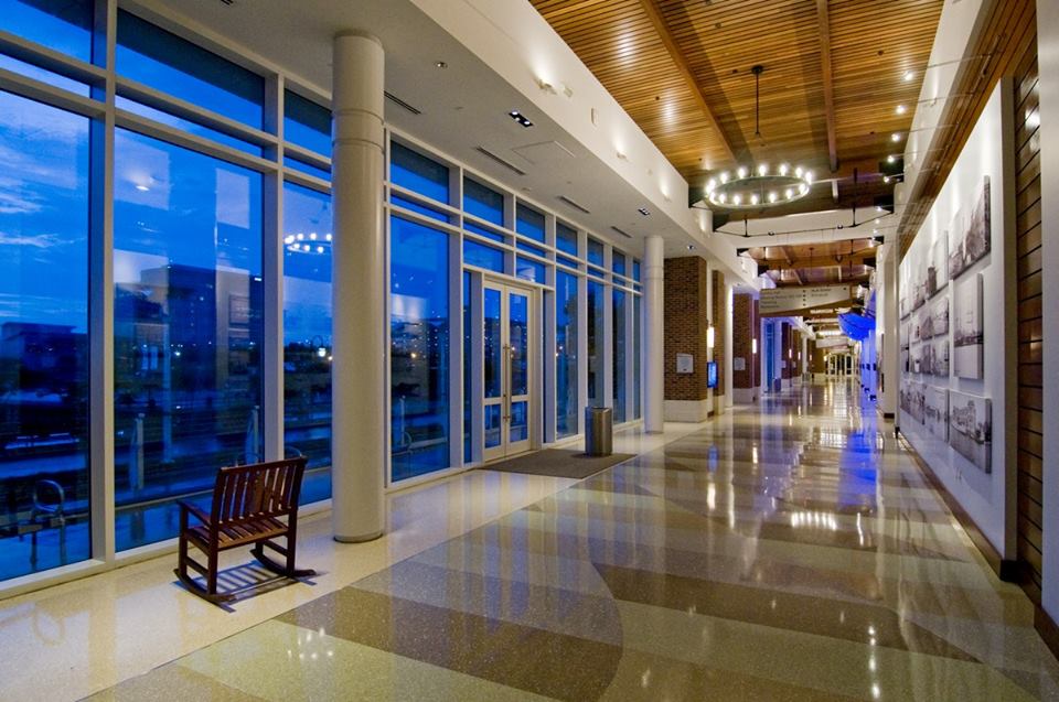 Interior corridor at dusk with floor-to-ceiling windows, wood ceiling accents, circular chandeliers, and historic photo displays along the wall.