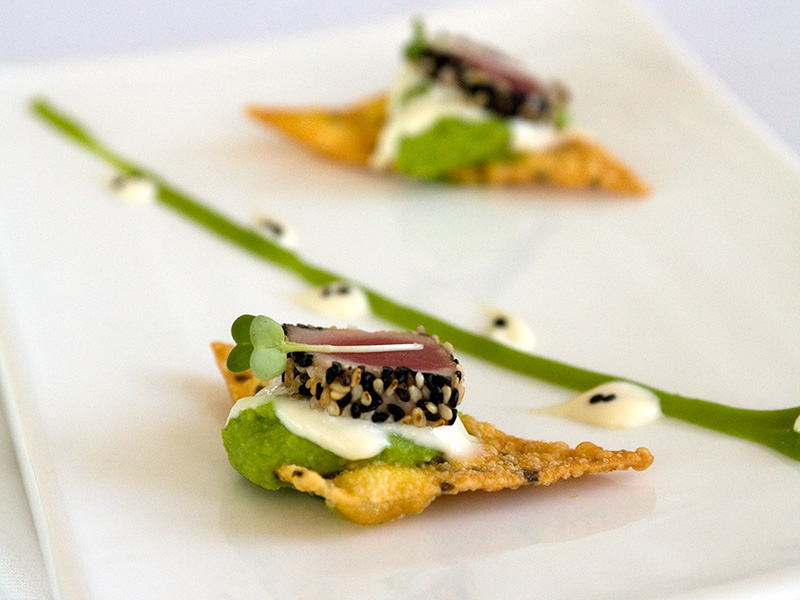 Plated hors d’oeuvre with two crisp wonton-style bites topped with green puree, white sauce, sesame-crusted tuna, and microgreens, arranged on a rectangular white plate.