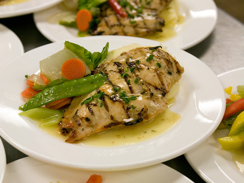 Plated grilled chicken breast with mixed vegetables and a light sauce, shown alongside other prepared plates in a kitchen setting.