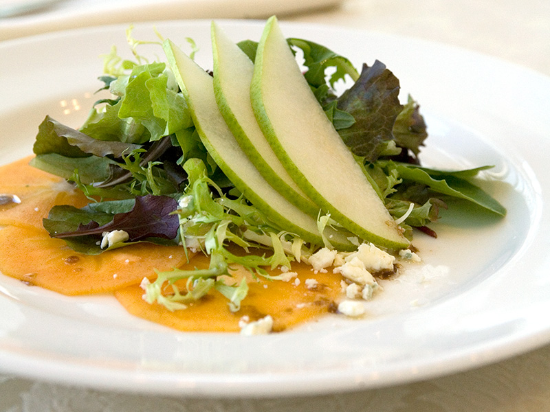 Plated salad with mixed greens, thin pear slices, crumbled cheese, and orange fruit slices on a white plate