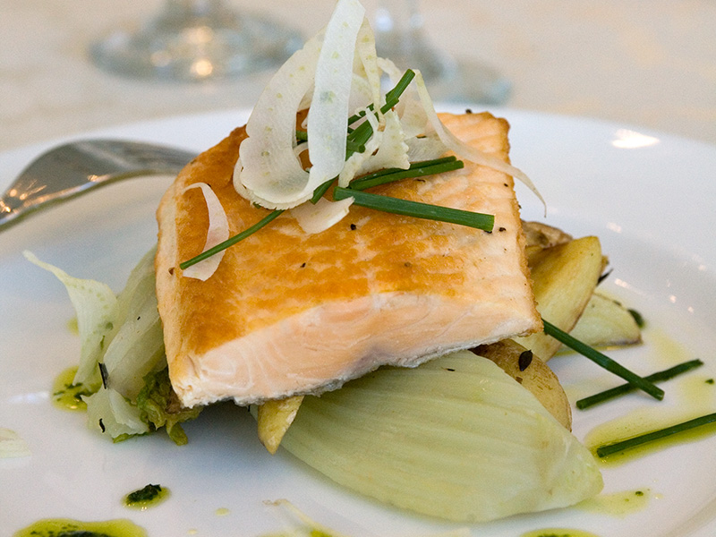 close-up of a plated salmon fillet served over vegetables and potatoes, topped with shaved fennel and chives
