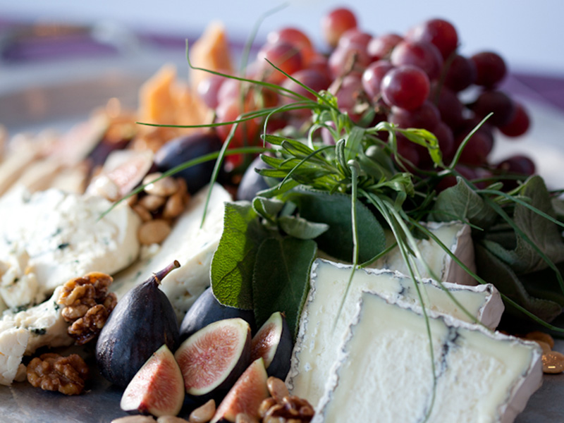 Close-up of a cheese board with soft cheeses, figs, red grapes, walnuts, pine nuts, and fresh herbs arranged on a tray.