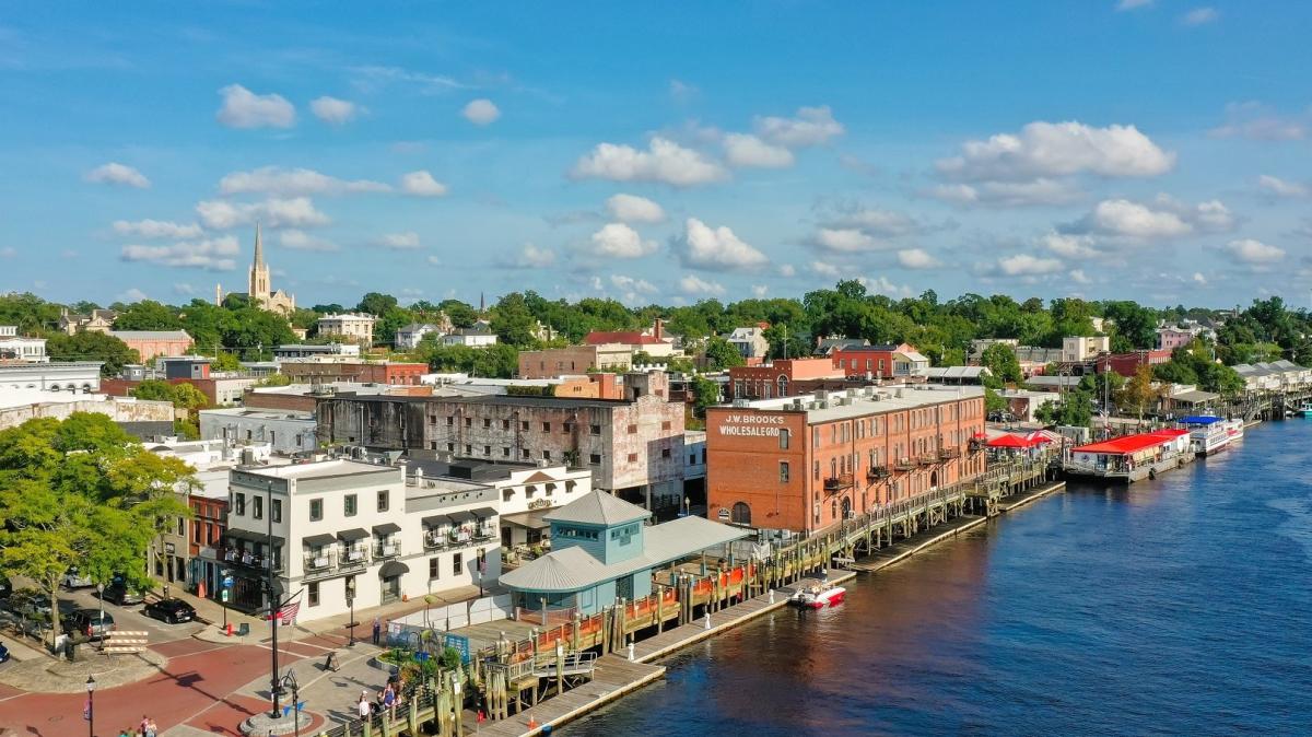 Aerial view of Wilmington’s downtown riverwalk and waterfront buildings along the river, with brick warehouses, docks, trees, and a church steeple under a bright blue sky.