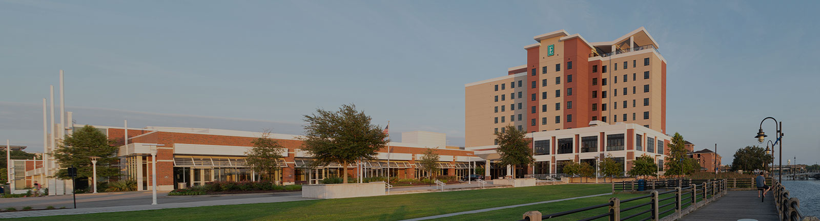 Waterfront view of the Wilmington Convention Center and adjacent hotel beside a riverwalk, with the river and marina extending along the right side under a clear sky.