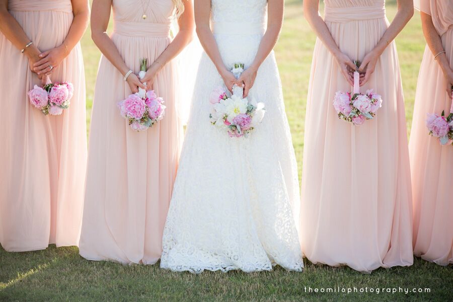 Close-up view of the bride and bridesmaids from the shoulders down, wearing blush pink and white gowns and holding small pink-and-white bouquets outdoors.