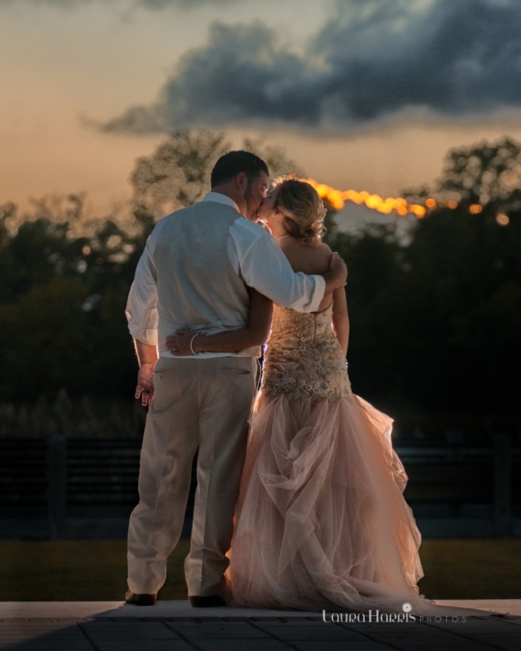 Bride and groom embracing and kissing at sunset, silhouetted against a dramatic sky with dark clouds and a glowing horizon.