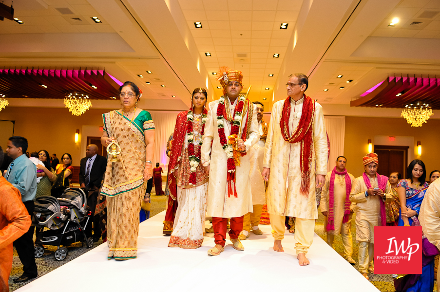 South Asian wedding entrance in a ballroom, with the groom in cream and red ceremonial attire walking down a white aisle alongside family members as guests watch from both sides.