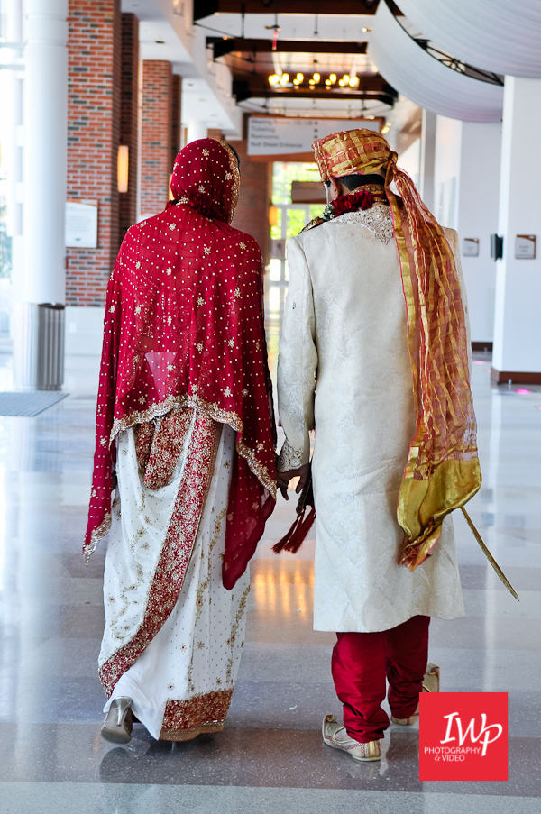 South Asian bride and groom walking hand in hand away from the camera through a bright interior corridor, wearing traditional red, gold, and cream wedding attire.
