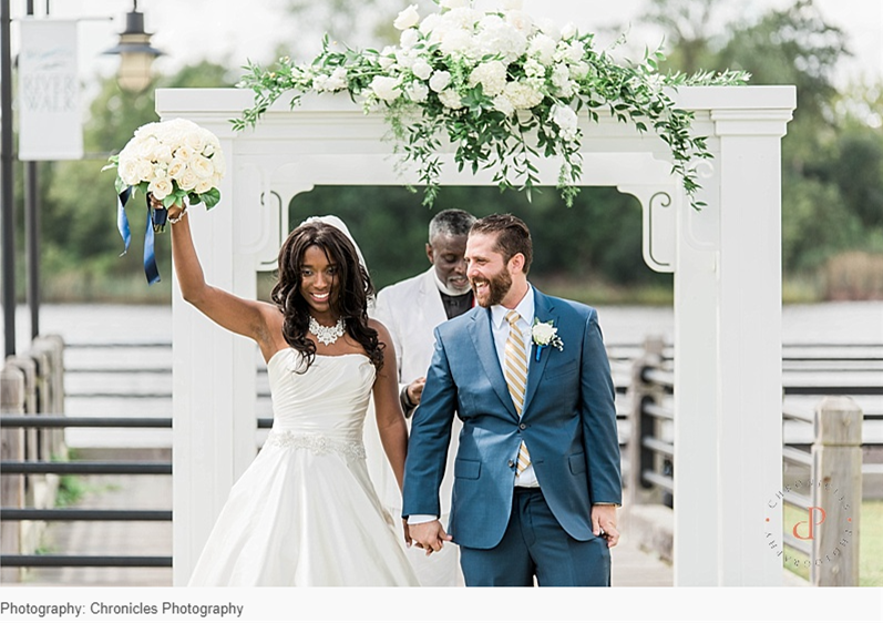 Bride and groom walking hand in hand beneath a white floral arbor by the waterfront, with the bride raising her bouquet as an officiant stands behind them.
