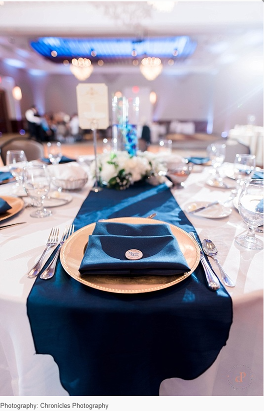 Elegant table setting at a ballroom event with a blue table runner, folded blue napkin on a gold charger, floral centerpiece, and softly blurred chandeliers in the background.