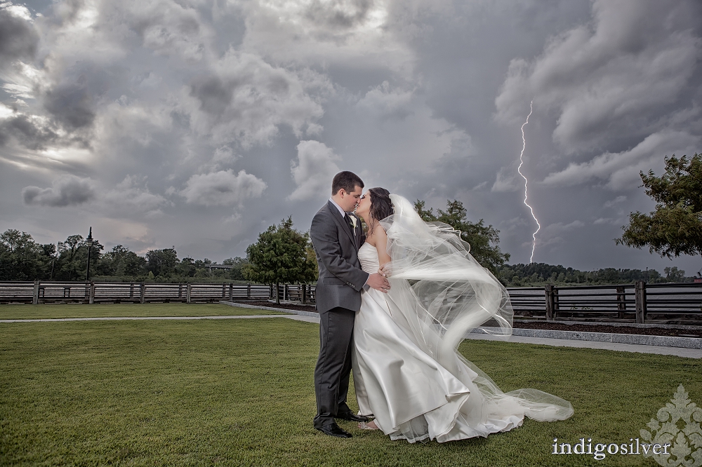 Bride and groom kissing on a waterfront lawn beneath dramatic storm clouds, with the bride’s veil blowing in the wind and a lightning strike visible in the background.