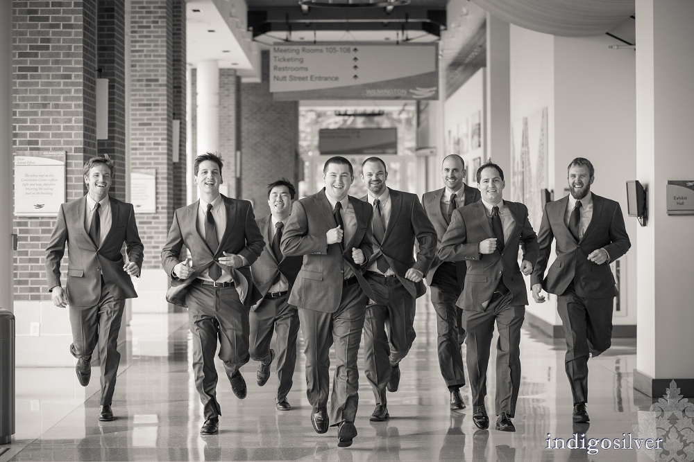 Black-and-white photo of seven men in matching suits running through a bright interior corridor, smiling as they move toward the camera.