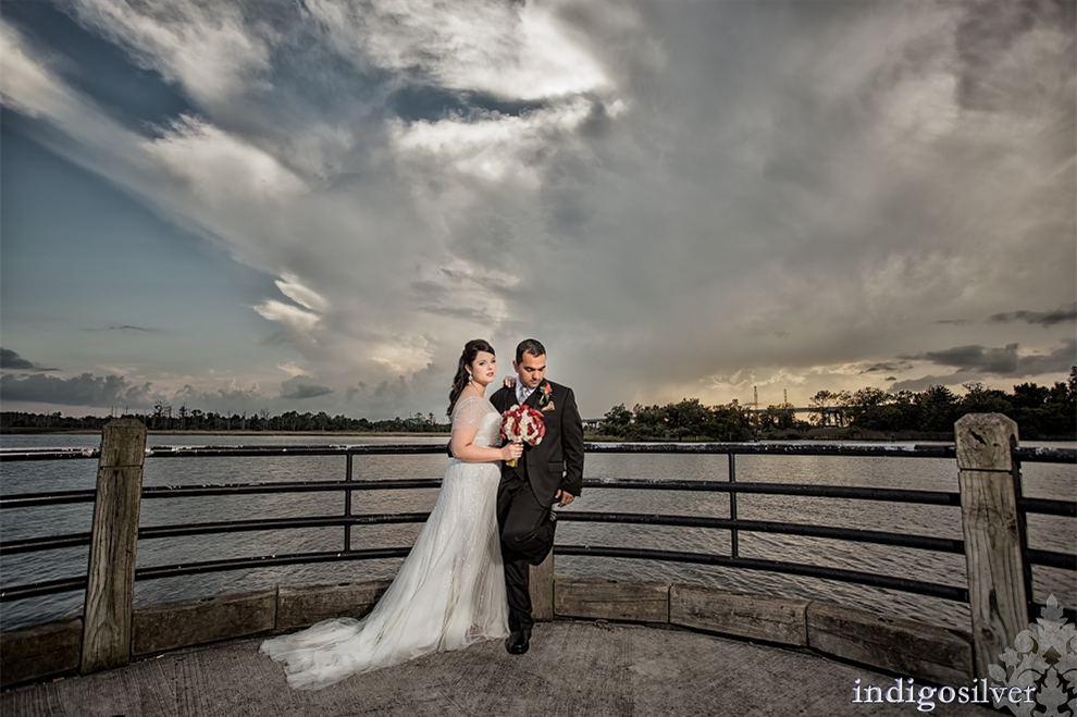 Bride and groom standing on a waterfront overlook, with the bride holding a bouquet and dramatic storm clouds filling the sky behind them.