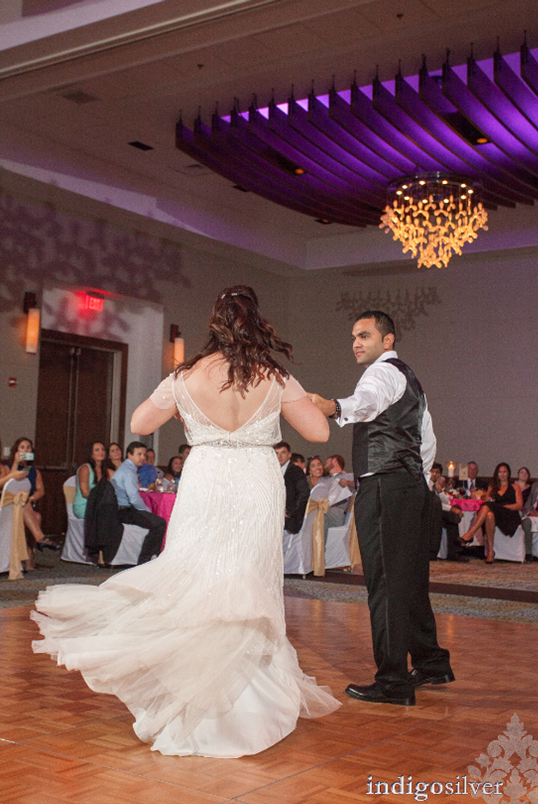 Bride and groom sharing a first dance in a ballroom, with guests seated around the dance floor beneath purple uplighting and chandeliers.