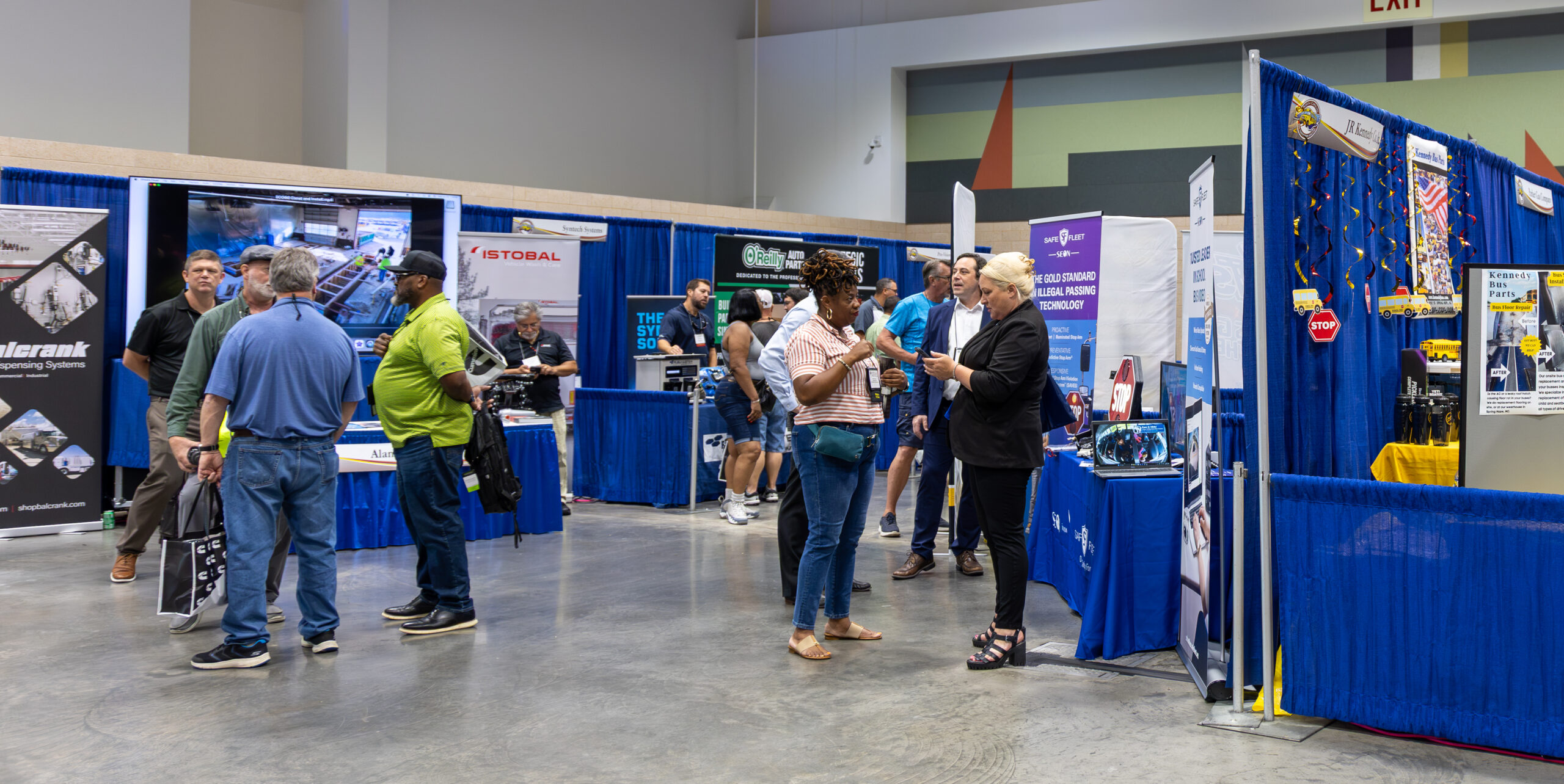 Trade show attendees talking and browsing exhibitor booths inside a convention hall, with blue drape displays, product signage, and demonstration screens.