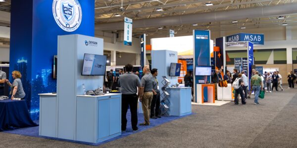Wide view of a convention center exhibit hall with large trade show booths, carpeted aisles, and attendees walking through the expo floor.