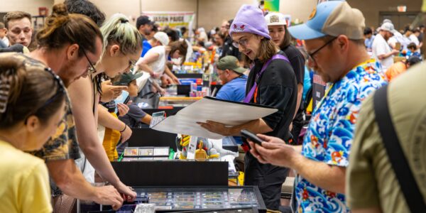 Crowded indoor expo or trading floor with attendees browsing display cases and tables, including one person holding a large framed item and another checking a phone.