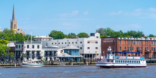 wilmington downtown Waterfront cityscape with historic brick buildings, a church steeple, a sailboat at the dock, and a white-and-blue riverboat cruising along the river on a clear day.