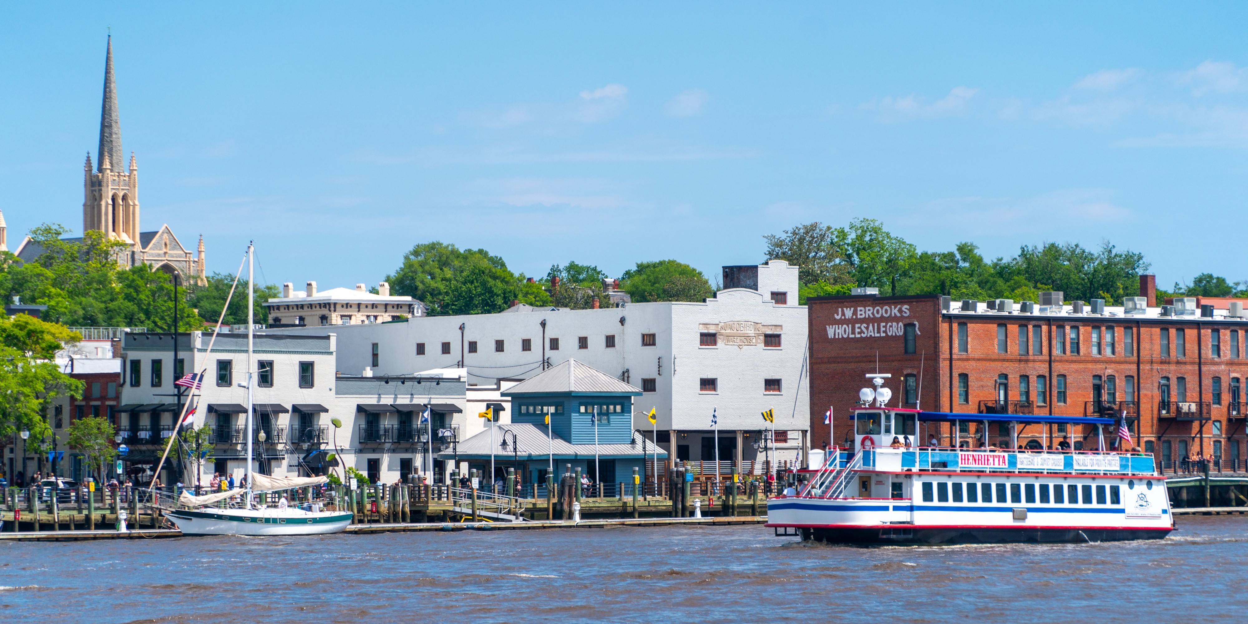 wilmington downtown Waterfront cityscape with historic brick buildings, a church steeple, a sailboat at the dock, and a white-and-blue riverboat cruising along the river on a clear day.