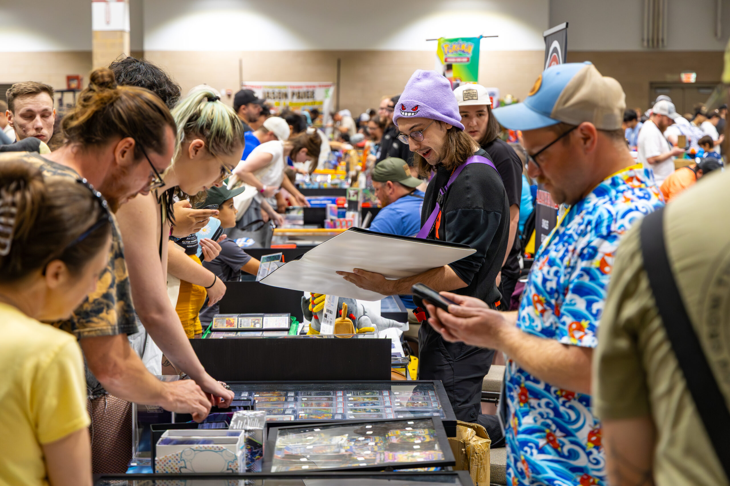 Crowded indoor expo or trading floor with attendees browsing display cases and tables, including one person holding a large framed item and another checking a phone.