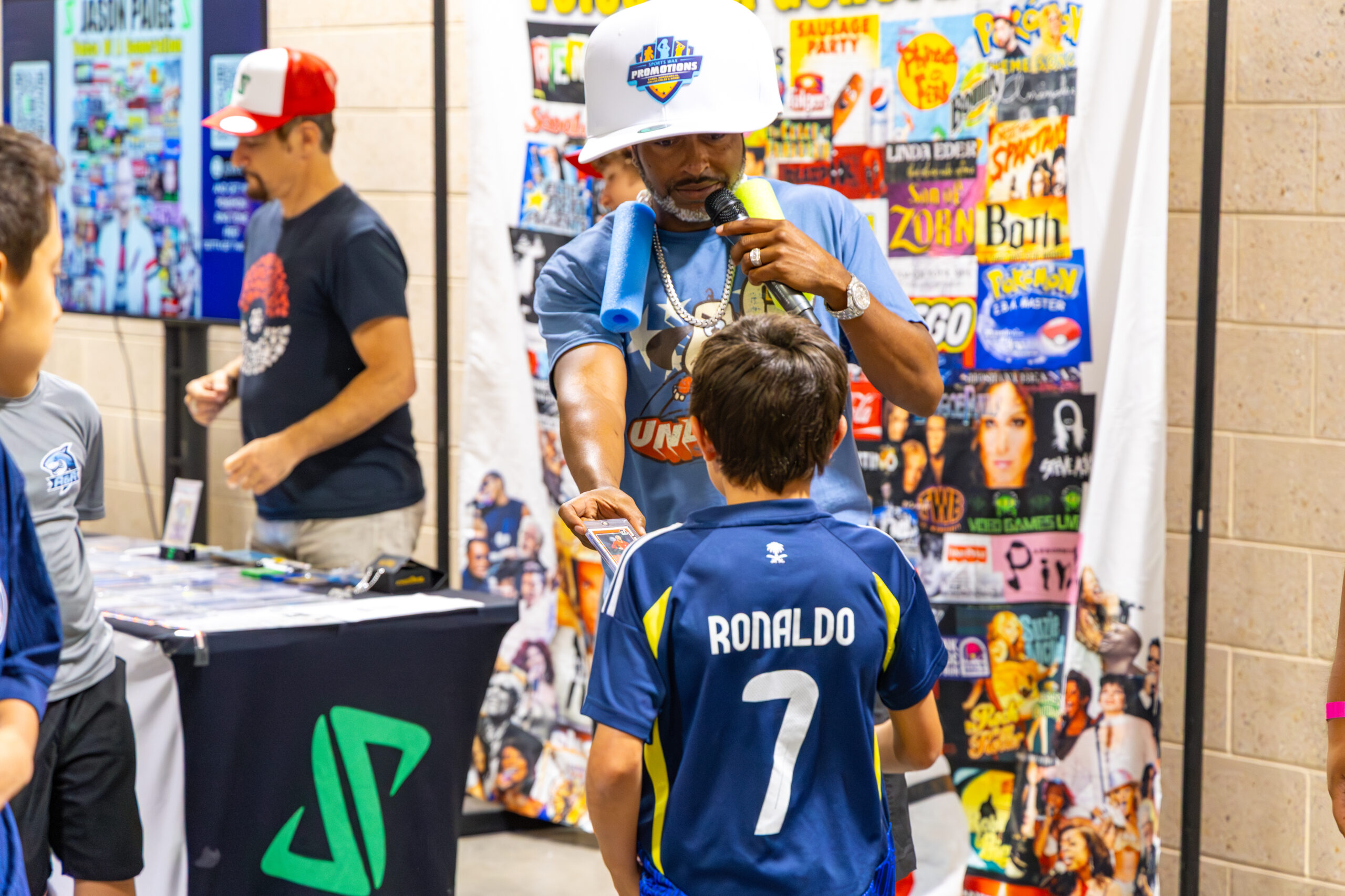 Man wearing a large white promotional hat and holding a microphone while handing a card to a child in a blue Ronaldo jersey at a busy convention event booth.