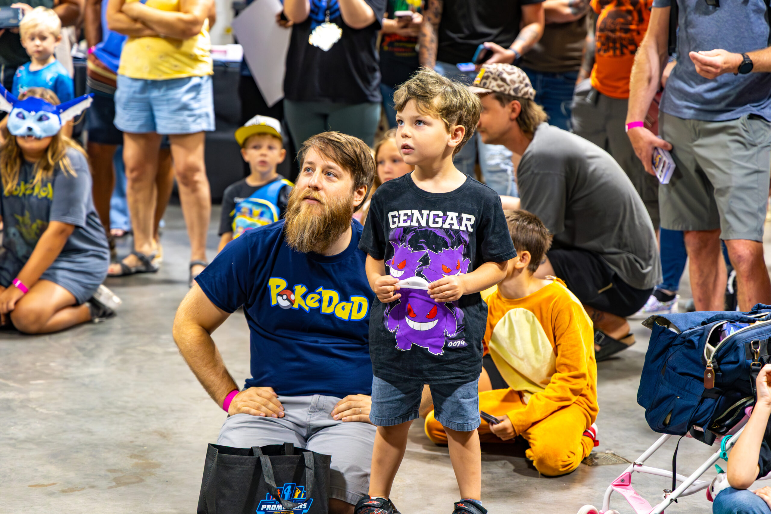 Child standing beside a seated adult in a crowded convention hall, with families gathered around and attendees watching an activity.