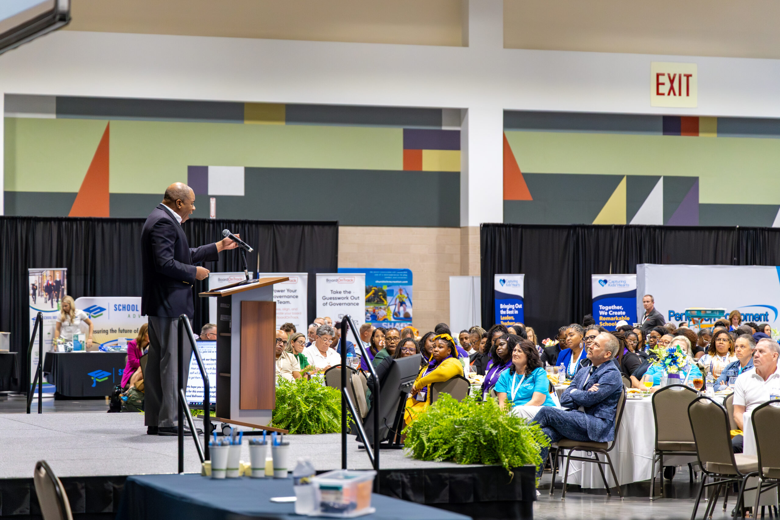 Man speaking at a podium during a convention hall event, with attendees seated at round tables and sponsor displays in the background.