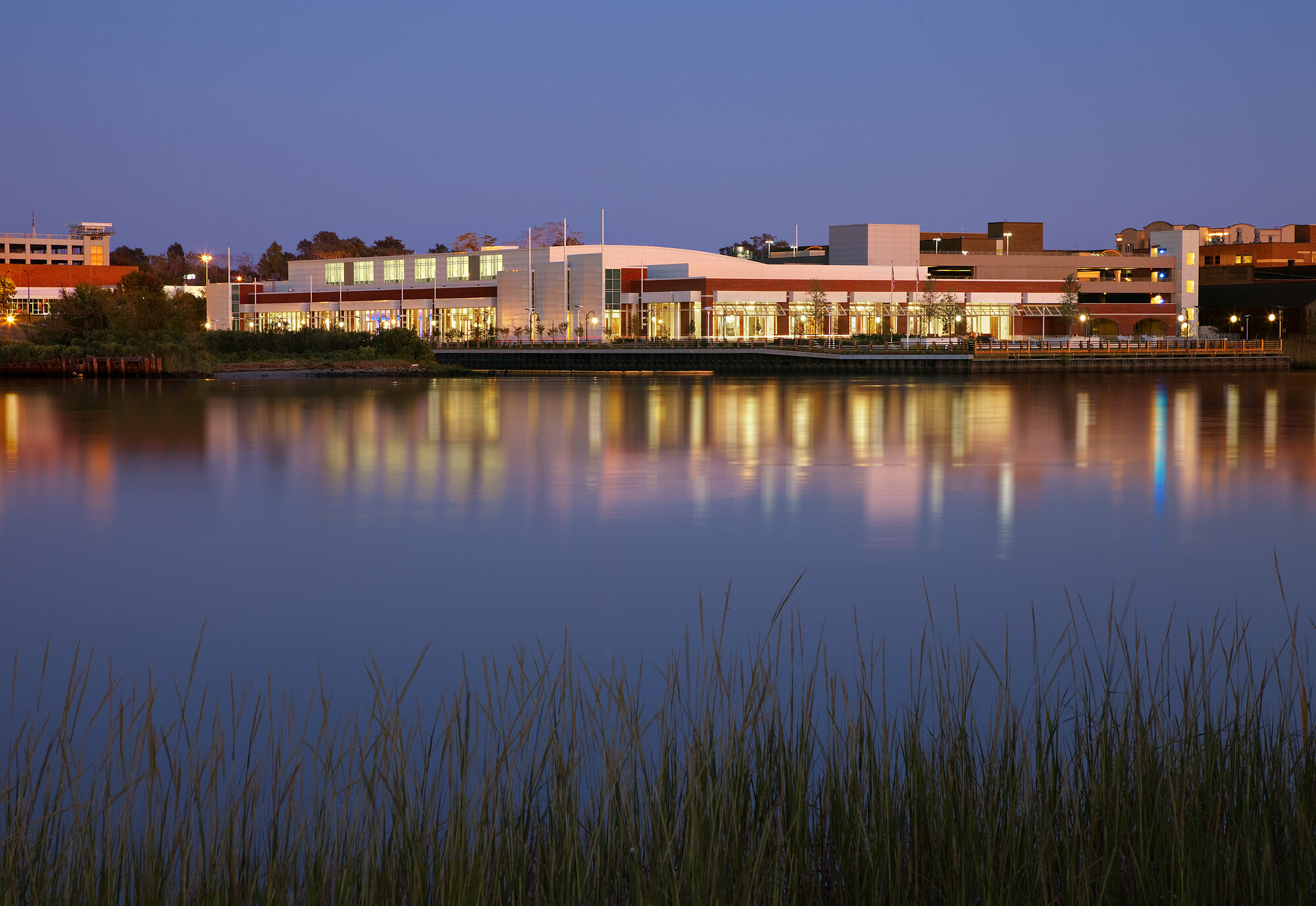 Twilight waterfront view of the Wilmington Convention Center with illuminated windows reflecting across calm water and marsh grass in the foreground.