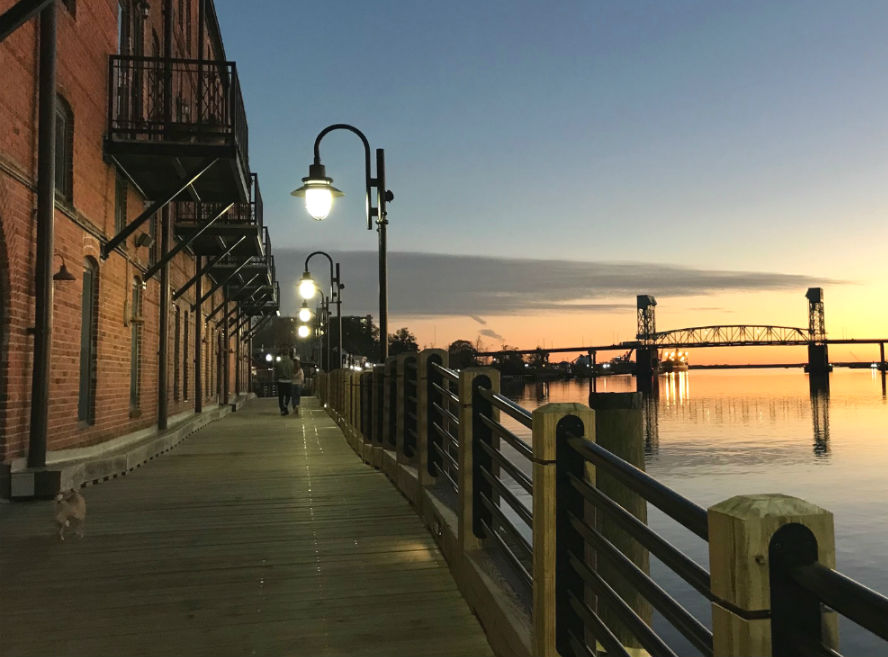 Riverwalk boardwalk at sunset beside a brick waterfront building, with glowing lampposts and a bridge spanning the river in the distance.