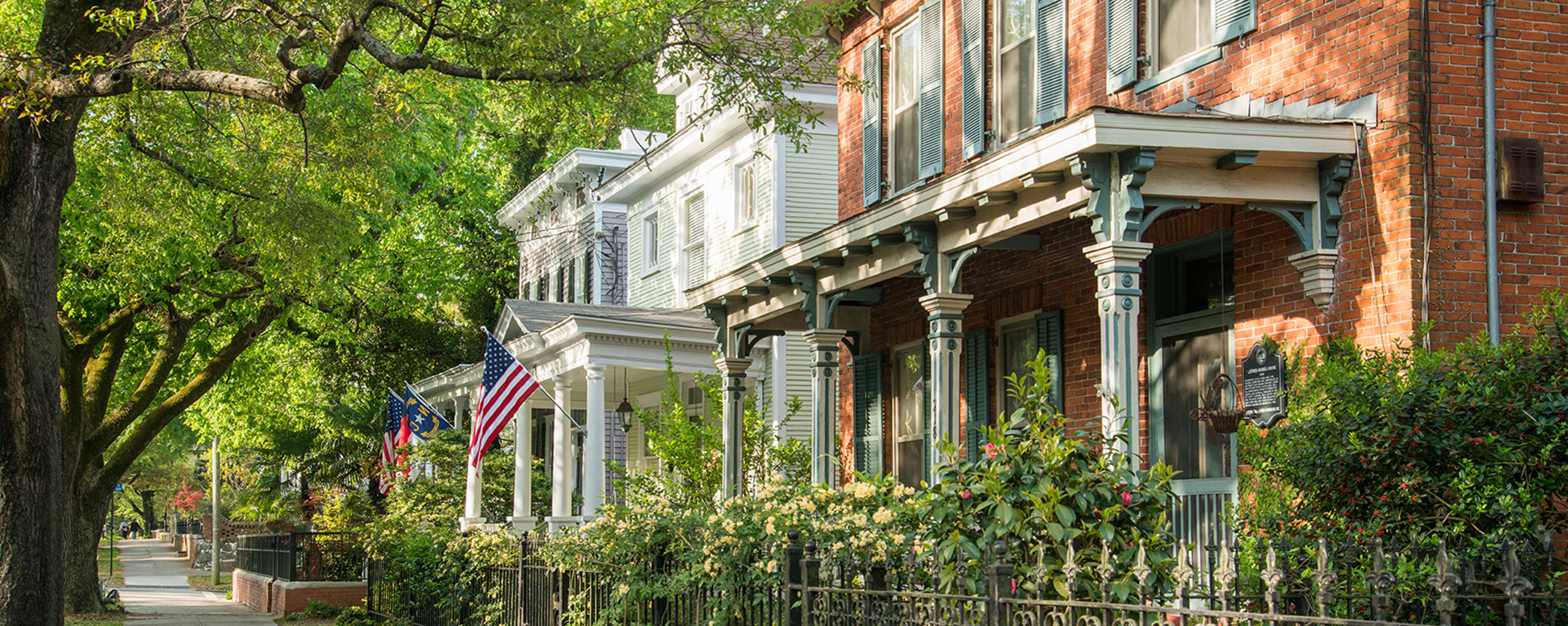 Tree-lined historic residential street with brick and white houses, front porches, wrought-iron fences, and American flags along a shaded sidewalk.